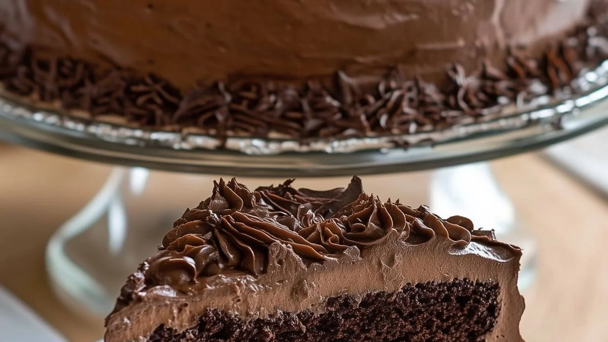 A Classic Chocolate Layer Cake with Chocolate Buttercream on a white stand, topped with chocolate shavings and served on a rustic wooden table